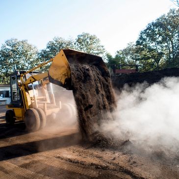 Yellow loader dumping dark soil with dust in a sunny outdoor area.