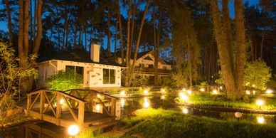 A warmly lit garden with a wooden bridge and house at night.