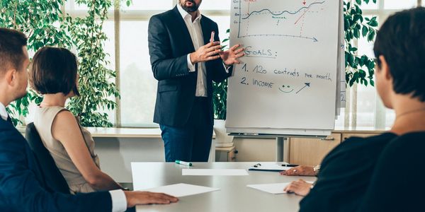 Man presents sales goals to colleagues using a flip chart in a bright office.