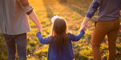 Child holding hands with two adults walking in a sunlit park.