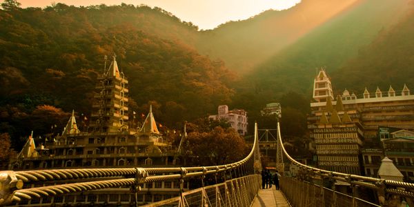 Sunlit suspension bridge leading to ornate temple buildings in a mountainous area.