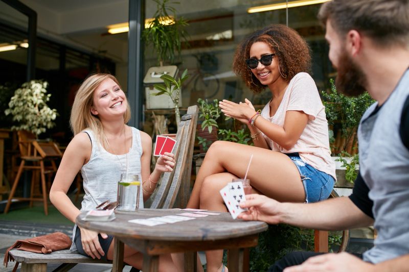 Three friends sitting in outdoor cafe and playing cards and having fun. Happy young people at sidewalk coffee shop enjoying playing poker game.