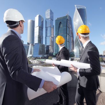 Engineers in hard hats reviewing blueprints with skyscrapers in the background.