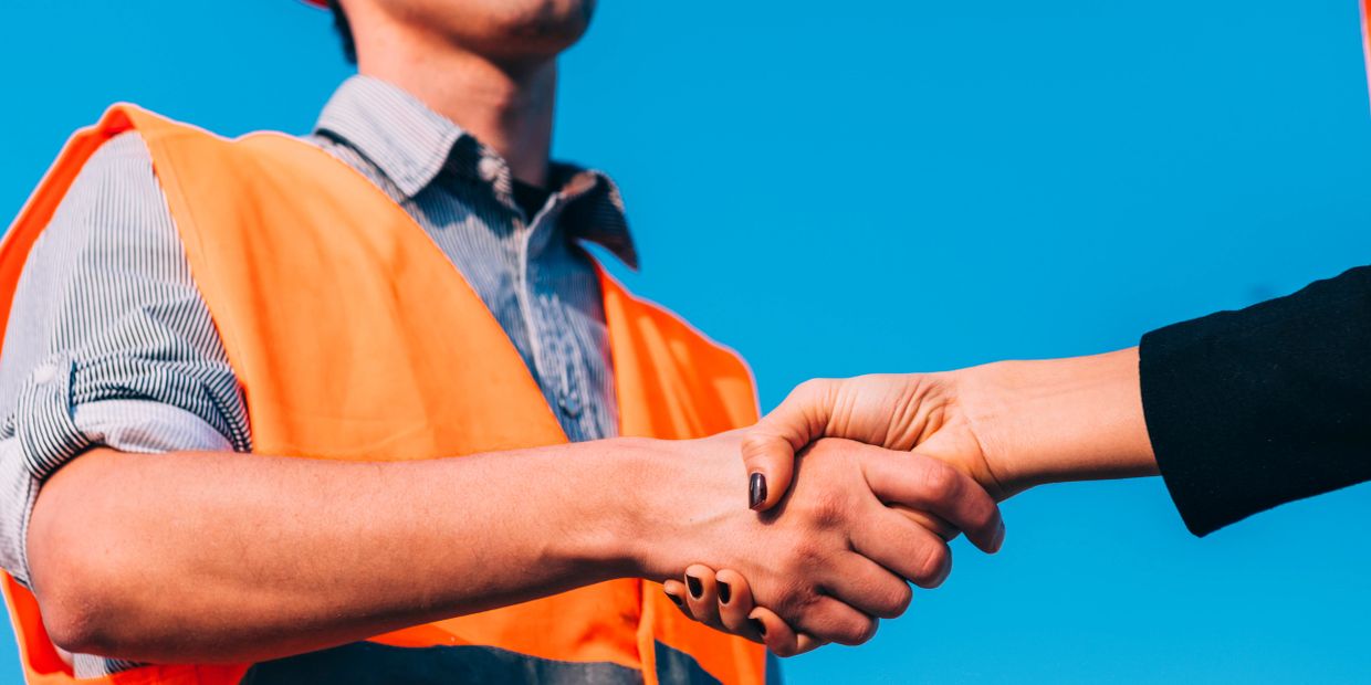 Handshake between a worker in an orange vest and another person against a clear blue sky.