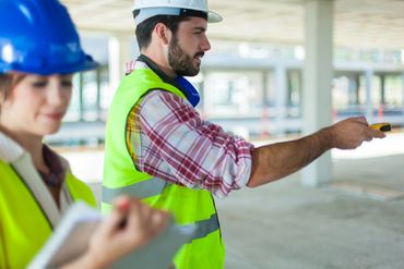 Two construction workers in safety gear discuss plans at a building site.