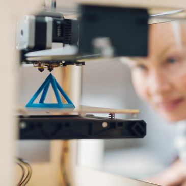 Woman observing a blue 3D-printed pyramid on a 3D printer.
