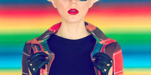 Fashionable woman with sunglasses, hat, and gloves against a colorful striped backdrop.