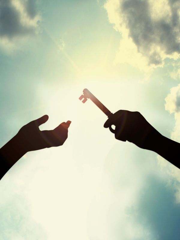 Silhouetted hands exchanging a vintage key against a bright sky with clouds.