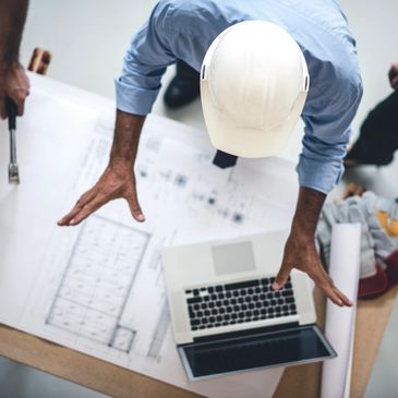 Engineer wearing a helmet reviews blueprints with a laptop on a table.