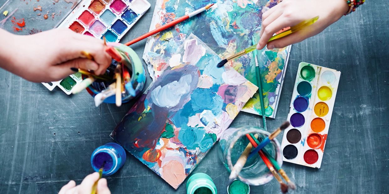 Hands holding paintbrushes over colorful palettes and paintings.