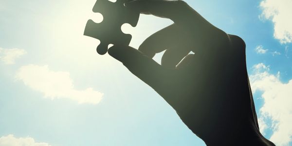 Hand holding a puzzle piece against a bright sky with clouds.