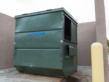 Large green dumpster with dented sides in an outdoor enclosure.