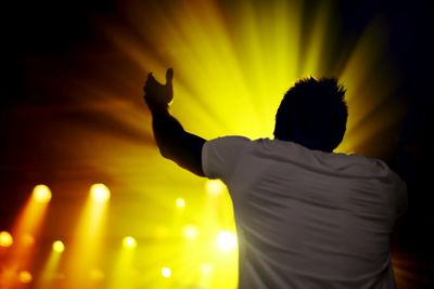 Person raising arms in celebration with bright yellow stage lights.