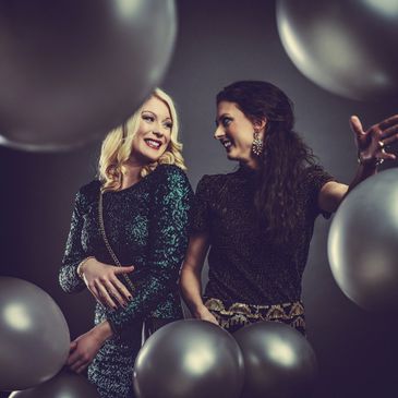 Two women joyfully posing among silver balloons in elegant dresses.