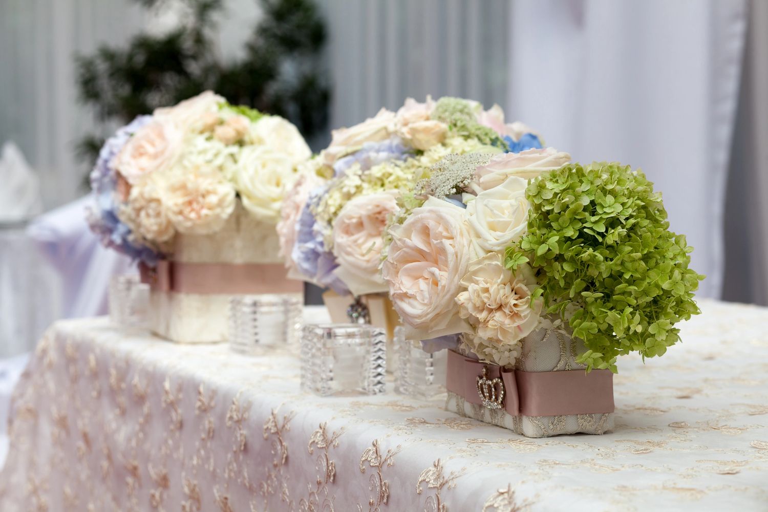 Elegant floral centerpieces with soft pastel roses and greenery on a lace-covered table.