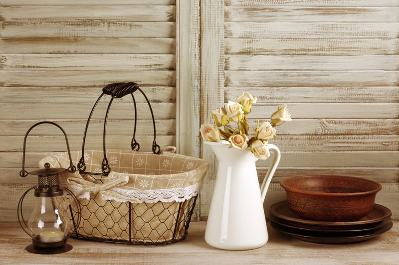 Rustic kitchen still life: wire basket, jug with roses bunch, ceramic dishware and lantern against vintage wooden shutters. Filtered toned image.