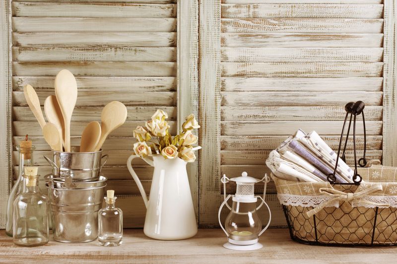 Rustic kitchen still life: wire basket with towels stack, galvanized buckets with wooden spoons, jug with roses bunch, glass bottles and lantern against vintage wooden shutters. Filtered toned image.
