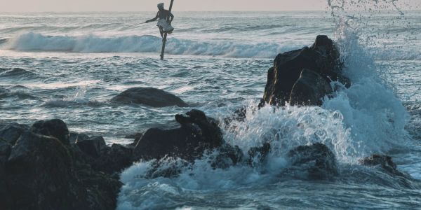A man fishing on a pole in the ocean during waves crashing on rocks.