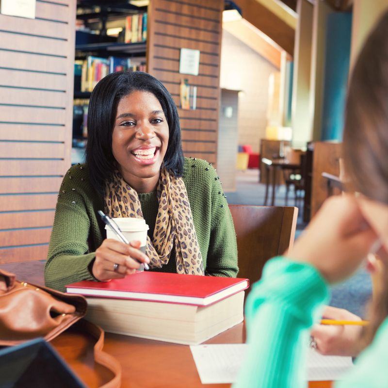 Teenage African American college age female student is smiling while sitting at desk in large modern library. College student is studying for exam and having coffee with study group of friends. She is wearing trendy casual clothing and a printed scarf.