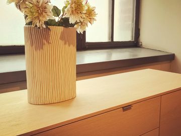 A textured vase with flowers on a wooden cabinet near a window.
