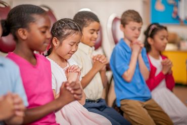 A group of children praying together with eyes closed and hands clasped.