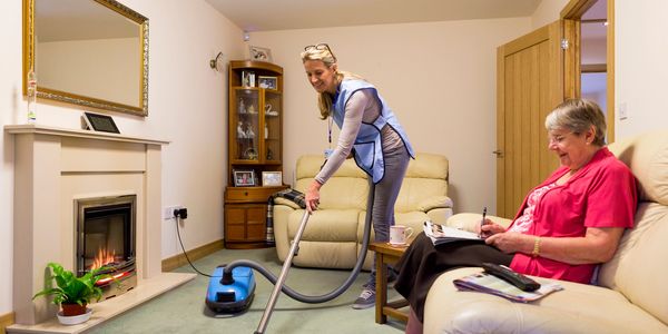 A caregiver vacuuming while an elderly woman writes on a notepad in a cozy living room.