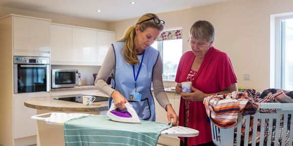 A caregiver irons clothes while an elderly woman watches.