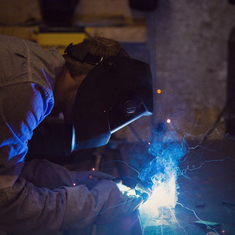 Young adult Caucasian man is working as a welder in a professional welding metal shop. Worker is wearing welder's mask and protective gloves and clothing. He is welding small pieces of metal together using a bright welding torch. Flames and sparks are coming from torch as it melts pieces of metal together on work table.