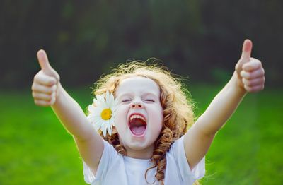 Joyful girl with a daisy in her hair giving two thumbs up outdoors.