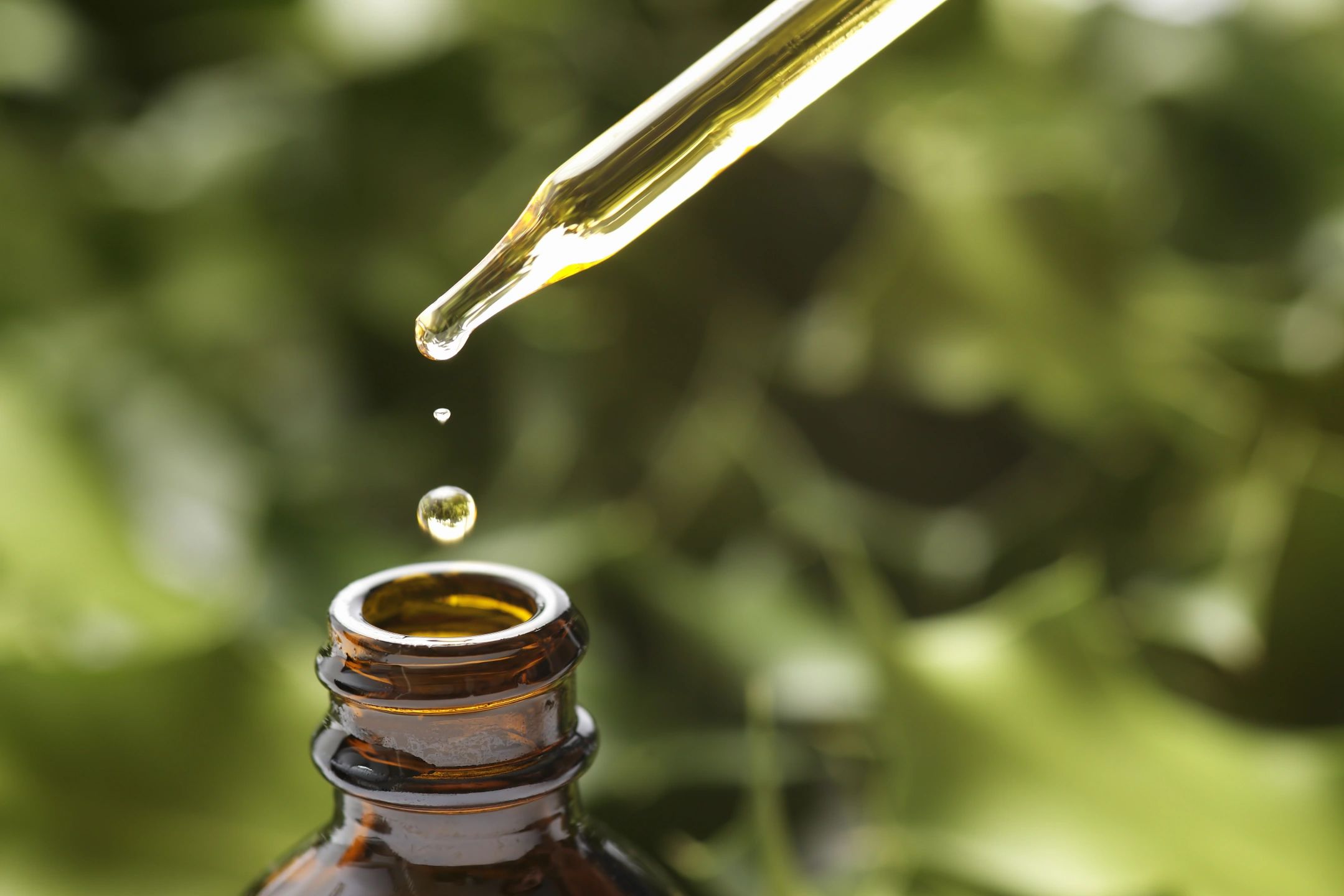 A dropper releasing oil into a brown bottle against a green leafy background.
