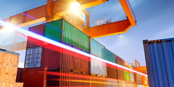Shipping containers stacked at a port under large orange cranes during sunset.