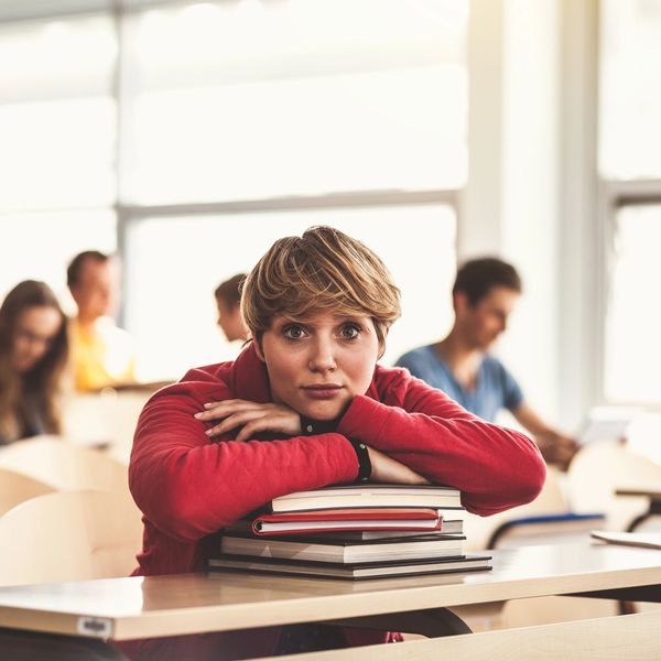 Student in red hoodie rests on books, looking thoughtfully at camera in classroom.