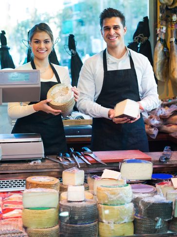Two smiling cheese shop workers presenting cheese behind the counter.