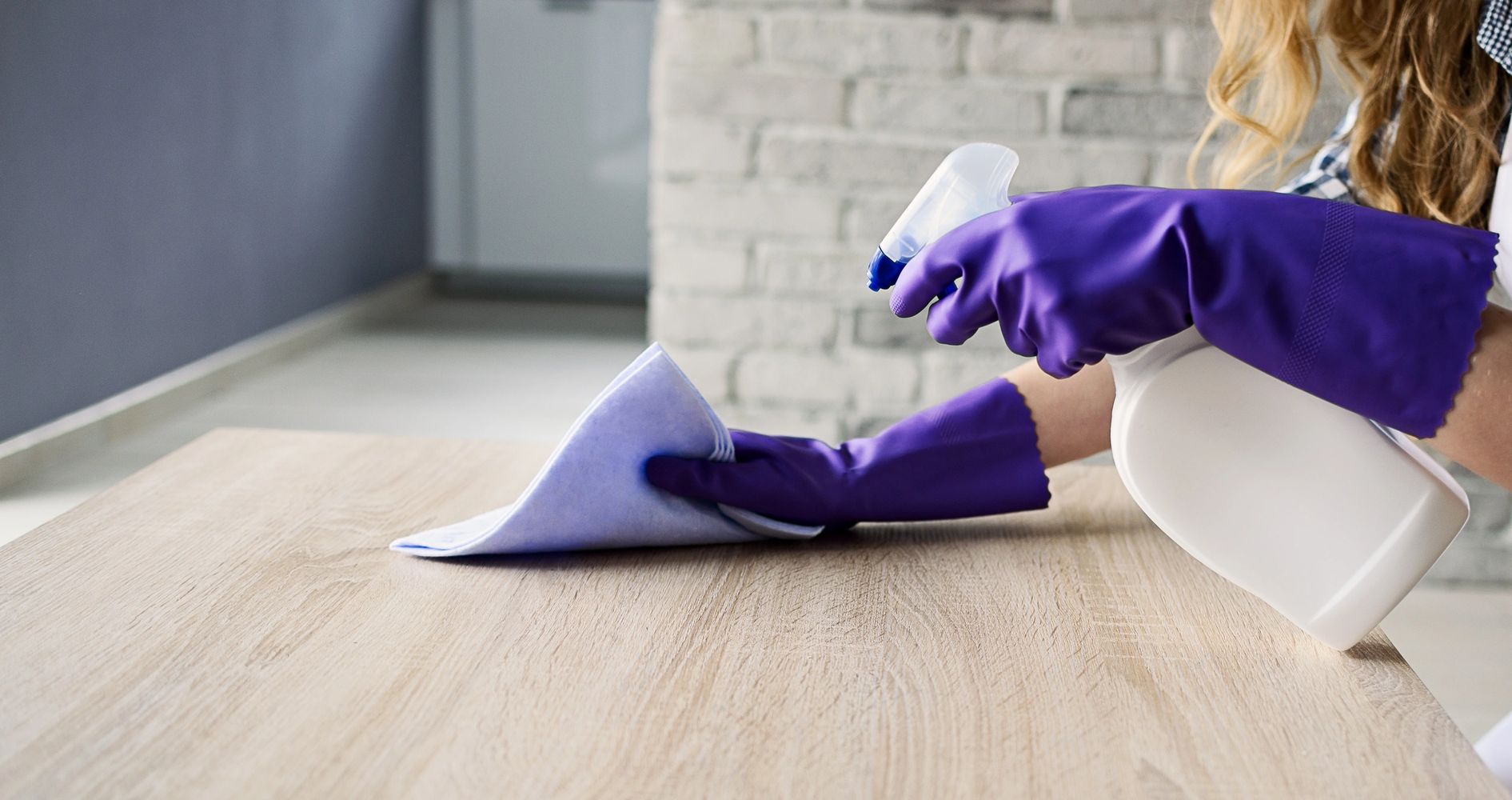 Person wearing purple gloves cleaning a wooden table with a spray bottle and cloth.