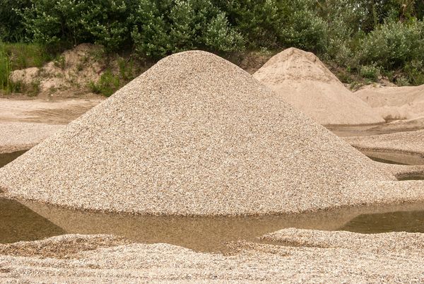 Large piles of gravel and sand near a water puddle with greenery in the background.