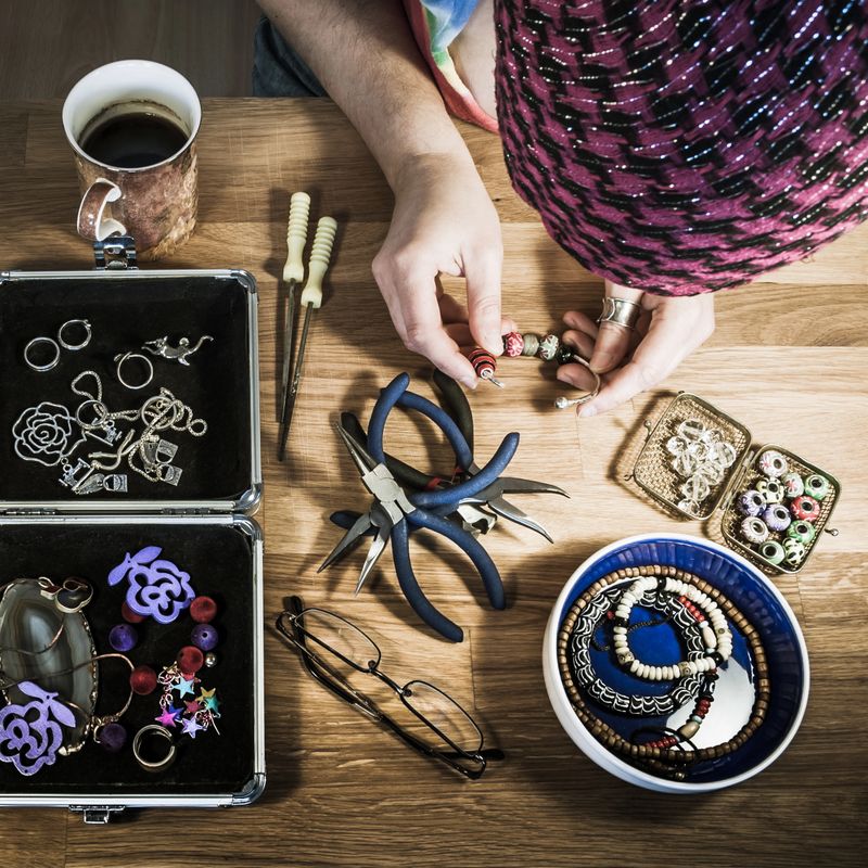 Woman making a bracelet on the wooden table using colorful beads, pliers and silver charms - square flatlay.