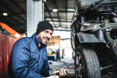 Mechanic in blue coveralls and beanie working on a car's wheel inside a garage.