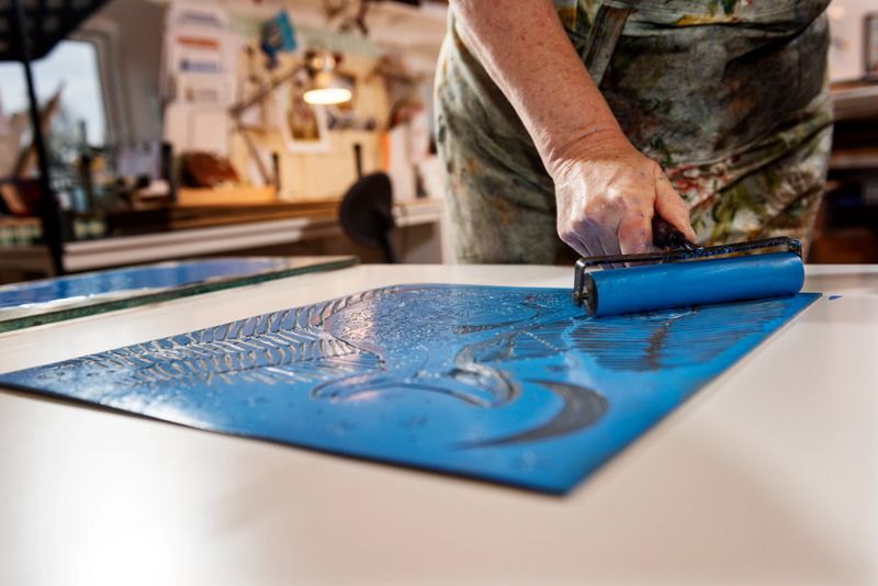 Portrait of a printmaker artist at work in her studio. She is rolling a  layer of blue ink onto her prepared lino cut prior to it being printed, subsequently different colours are added until the print is finished. Horizontal format with a tight composition with the the point of focus  on her hand as she grips the roller giving a nice out of focus background. Photographed at a her artists studio on the island of Møn Denmark.