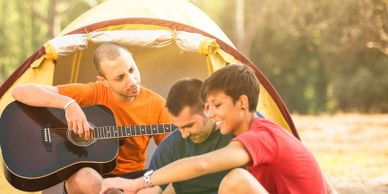 Three friends enjoying music and camping outdoors near a tent.