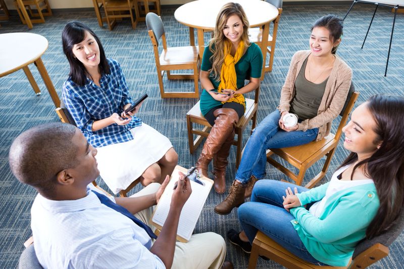 Diverse young creative professionals discuss project status for client in discussion group. A mid adult African American man is leading the group and giving assignments.  They are sitting in chairs in a circle. They are attentively listening to the project manager.
