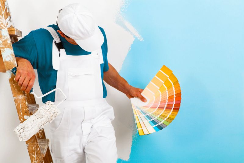 portrait of painter man with paintroller and wooden vintage ladder showing a color palette, on half painted wall