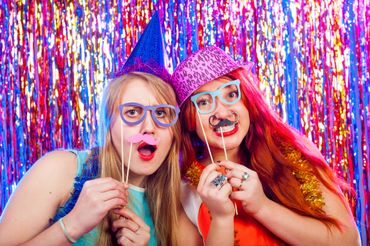Two women at a colorful party holding funny glasses and mustache props.