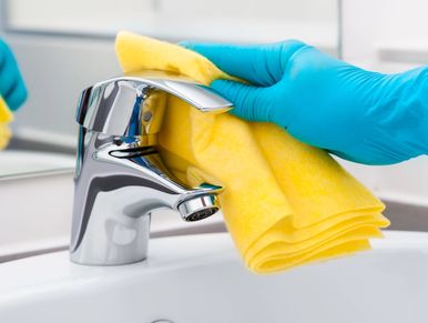 Hand wearing blue glove cleaning a bathroom faucet with yellow cloth.