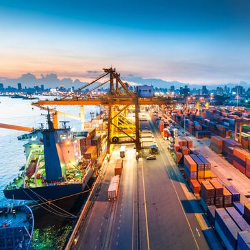 Container ship docked at a bustling port with cranes and stacked containers at dusk.