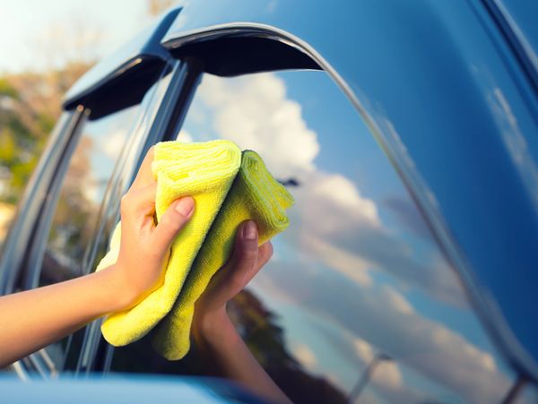 Person cleaning a car window with a yellow cloth on a sunny day.