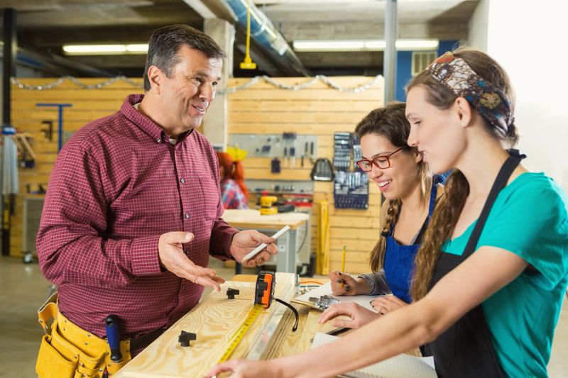 Young female apprentices listent to mid adult manager explain the details of their project for a client. The women are using a tape measure to measure a piece of wood. The man gestures as he talks. The man is wearing a yellow tool belt. Caucasian woman is wearing a scarf in her hair and is wearing a black apron. Hispanic woman is wearing a blue apron and red glasses. Tools are hanging on the wall in the background.