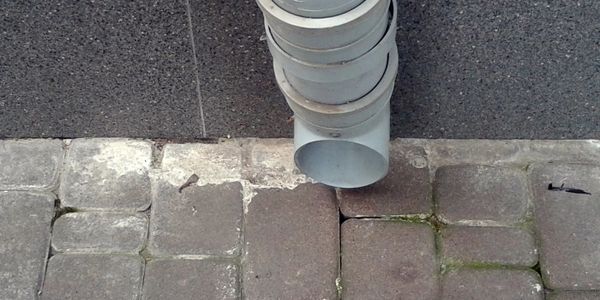 Gray drainage pipe extending from a wall over stone pavement.