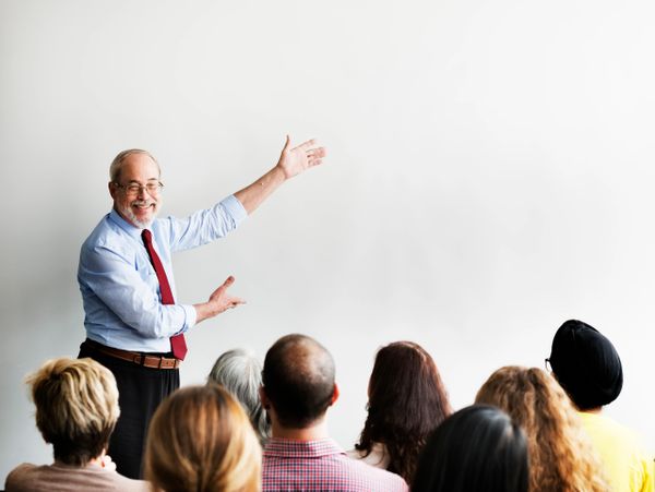 Older man giving a presentation to an audience in a bright room.
