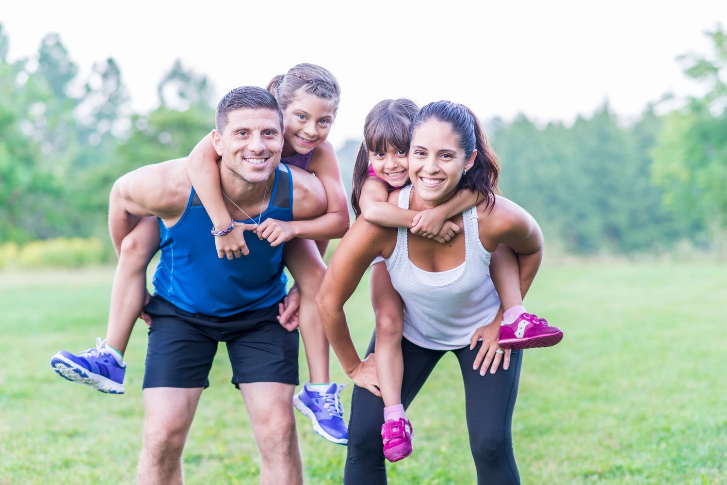 Smiling parents outdoors with two young children on their backs, representing active family wellness