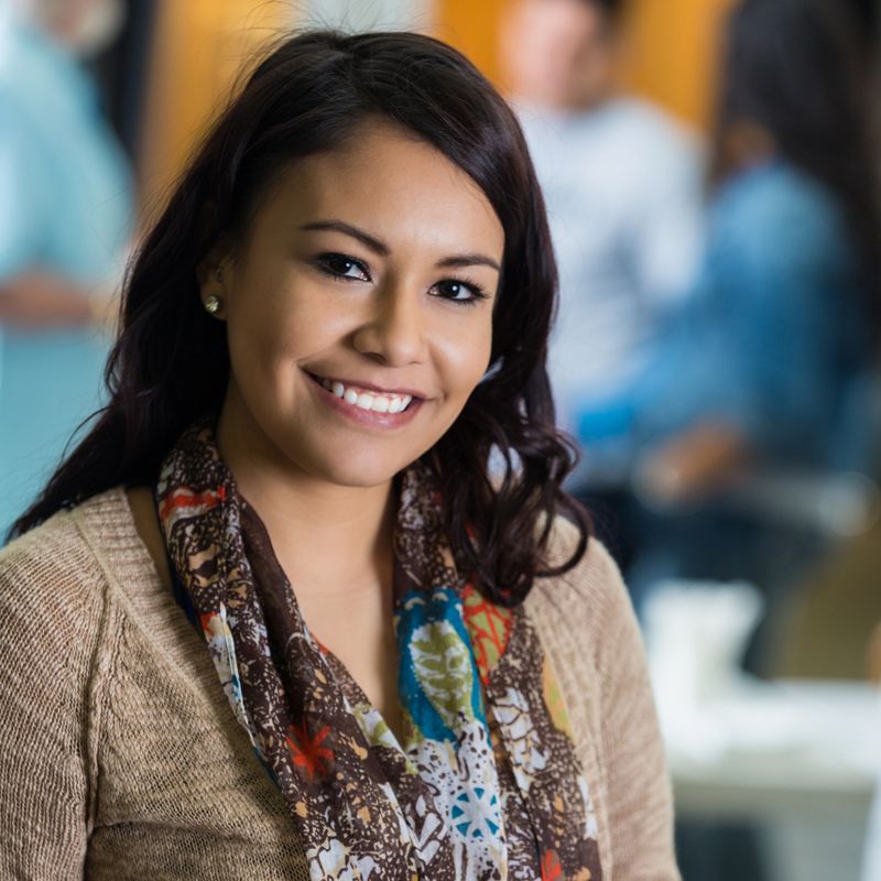 Pretty young Hispanic female volunteer at local soup kitchen or food drive. She has brown eyes and brown hair. She is wearing a tan sweater and has patterned scarf around her neck. She is smiling at the camera. Volunteers are working in the background.